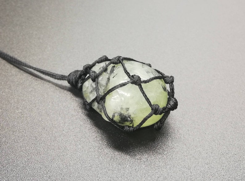 May include: A green and black crystal stone pendant is wrapped in a black macrame cord. The pendant is tied to a black cord.