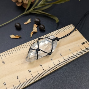 May include: A clear quartz crystal wrapped in black string, hanging from a black cord. The crystal is approximately 4 cm long and is resting on a wooden ruler with centimetre markings.