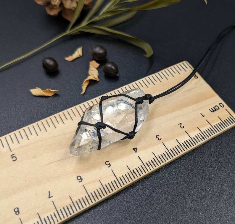 May include: A clear quartz crystal wrapped in black string, hanging from a black cord. The crystal is approximately 4 cm long and is resting on a wooden ruler with centimetre markings.
