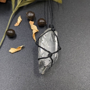 May include: A clear quartz crystal wrapped in black cord, hanging from a black cord necklace. The crystal is a point shape and is tied with a macrame knot.