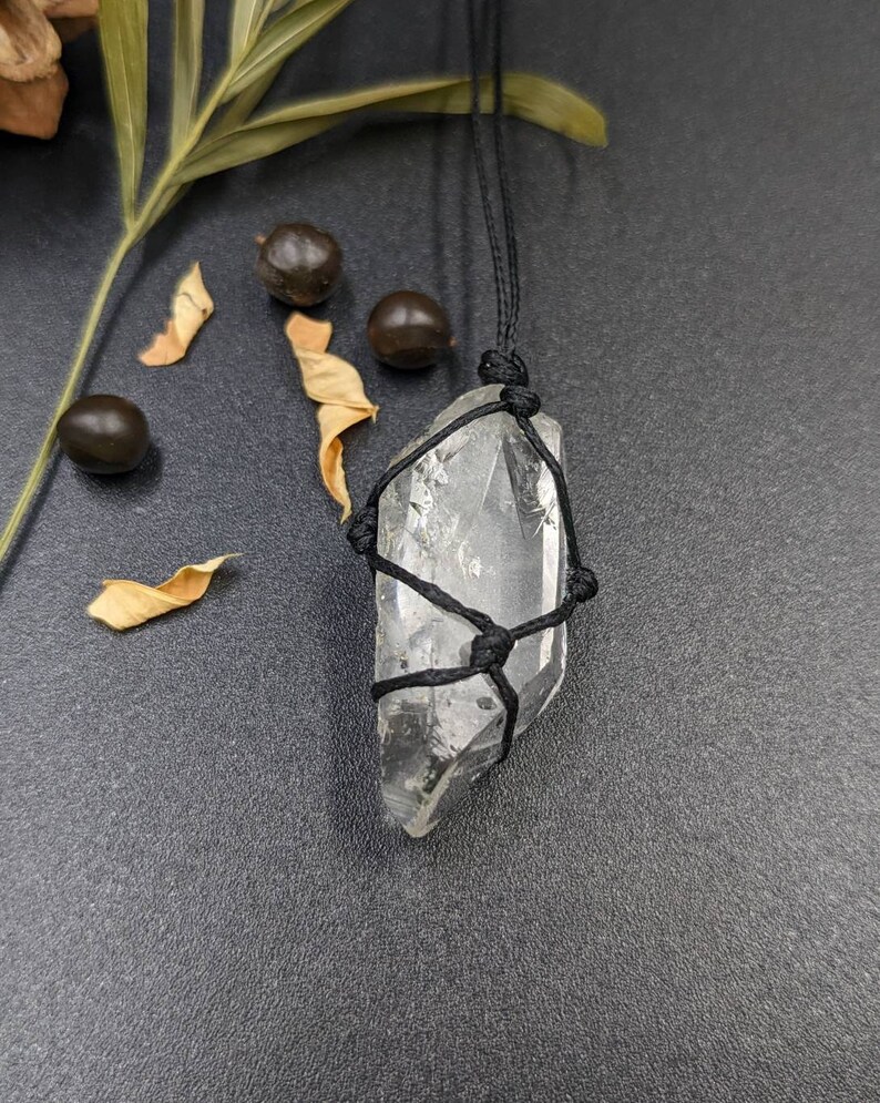 May include: A clear quartz crystal wrapped in black cord, hanging from a black cord necklace. The crystal is a point shape and is tied with a macrame knot.