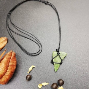 Pu&ograve; includere: Un ciondolo di pietra verde a forma triangolare appeso a una collana di cordoncino nero. Il ciondolo &egrave; legato al cordoncino con un nodo. La collana &egrave; su una superficie grigio scuro con foglie marroni e piccoli semi.