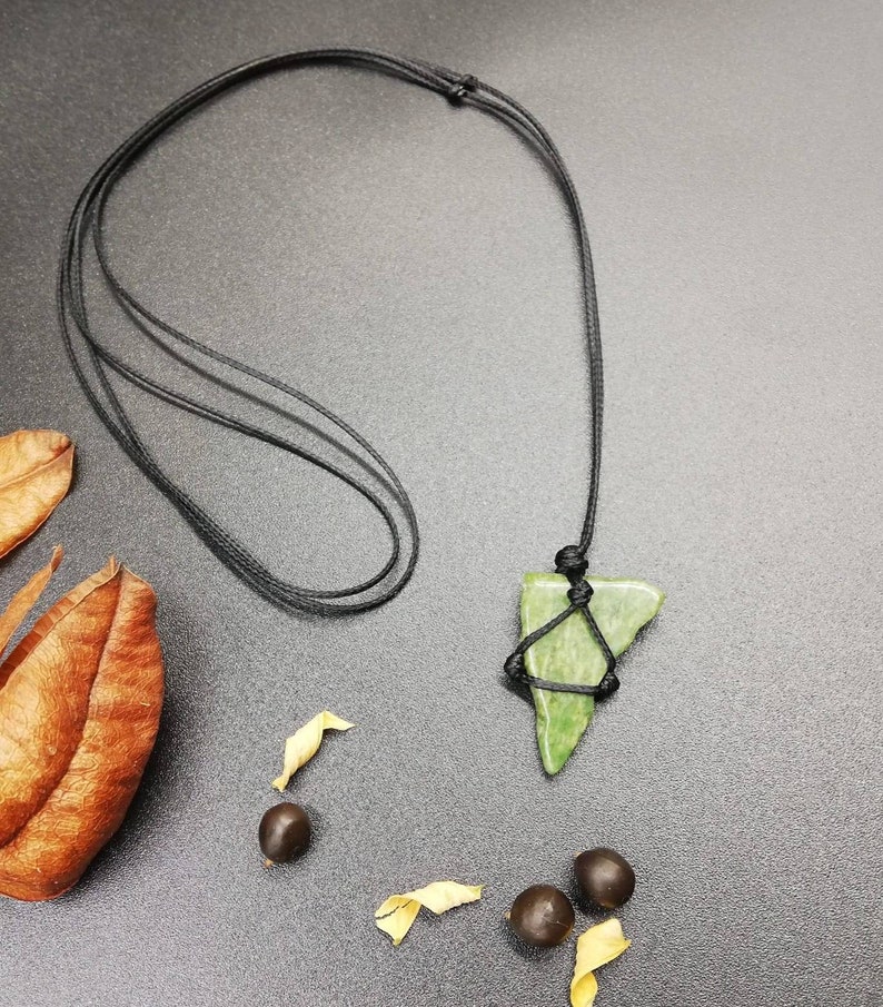 Pu&ograve; includere: Un ciondolo di pietra verde a forma triangolare appeso a una collana di cordoncino nero. Il ciondolo &egrave; legato al cordoncino con un nodo. La collana &egrave; su una superficie grigio scuro con foglie marroni e piccoli semi.