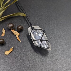 May include: A clear quartz crystal wrapped in black cord, hanging from a black cord necklace. The crystal is a point shape and has a natural, raw look.