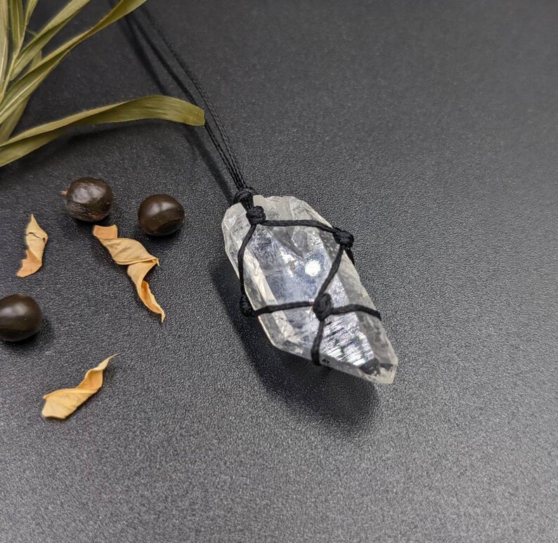 May include: A clear quartz crystal wrapped in black cord, hanging from a black cord necklace. The crystal is a point shape and has a natural, raw look.