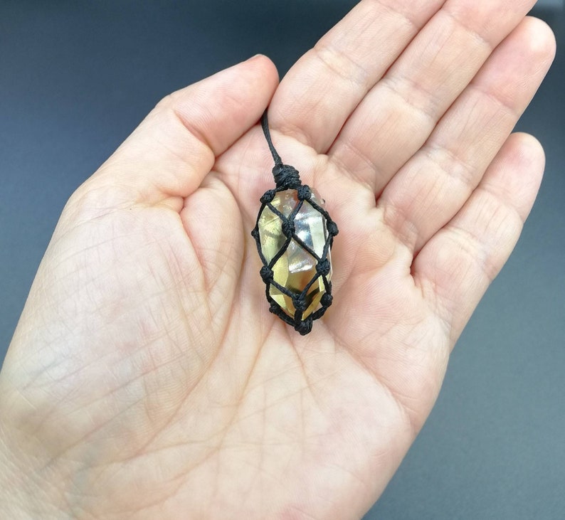 May include: A yellow crystal pendant in a black macrame knot. The crystal is faceted and has a natural, raw look. The pendant is hanging from a black cord.