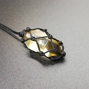 May include: A clear, faceted, yellow crystal is encased in a black macrame knotting. The crystal is suspended from a black cord.