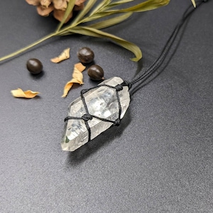 May include: A clear quartz crystal wrapped in black macrame cord, hanging from a black cord necklace. The crystal is a point shape and has a natural, raw look.
