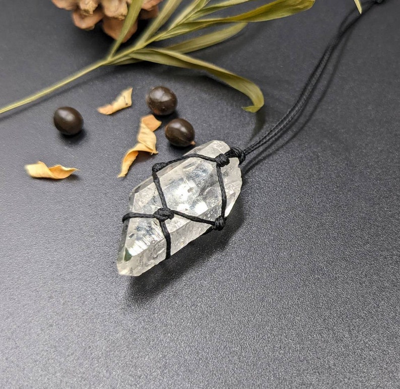May include: A clear quartz crystal wrapped in black macrame cord, hanging from a black cord necklace. The crystal is a point shape and has a natural, raw look.