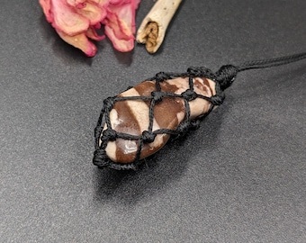 Op de afbeelding: Een bruin en wit gestreepte steen is gewikkeld in een zwart macramékoord, waardoor een hanger ontstaat. Het koord heeft een lus aan de bovenkant om op te hangen.