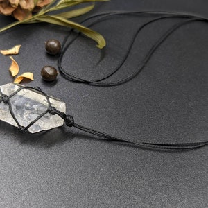 May include: A clear quartz crystal pendant wrapped in black cord, hanging from a black cord necklace. The pendant is tied with a macrame knot.