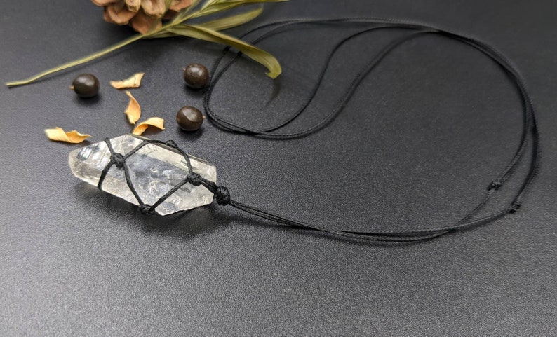 May include: A clear quartz crystal pendant wrapped in black cord, hanging from a black cord necklace. The pendant is tied with a macrame knot.