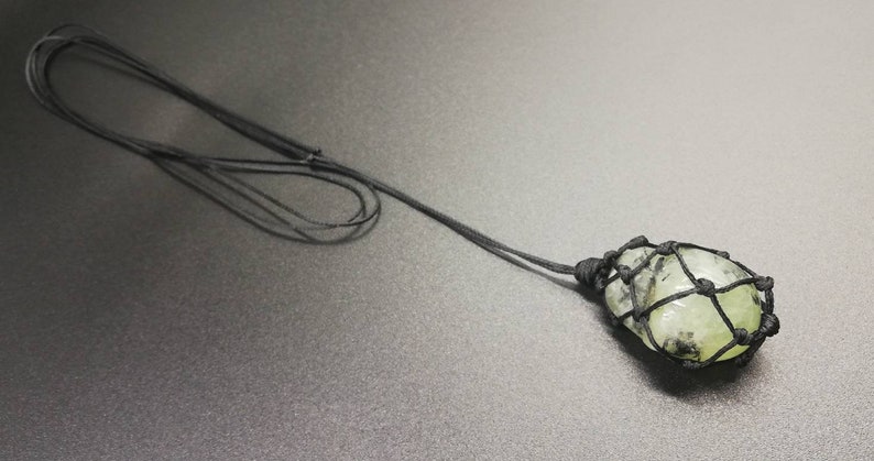 May include: A green stone pendant with black string knotted around it, hanging from a black cord.