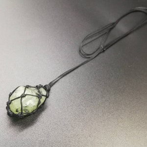 May include: A green crystal stone wrapped in black cord, hanging from a black cord necklace.