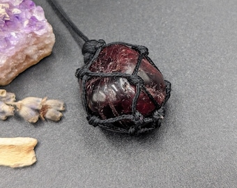 May include: A dark red gemstone pendant with a black macrame cord. The gemstone is wrapped in a black cord and has a natural, rough texture.
