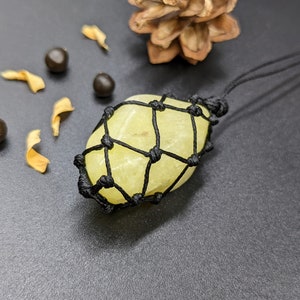 Può includere: Una pietra di cristallo gialla avvolta in una rete di macramè nera. La pietra è legata con un cordoncino nero.