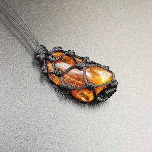 Può includere: Una pietra di ambra scura avvolta in un cordoncino nero annodato, creando una collana pendente annodata.