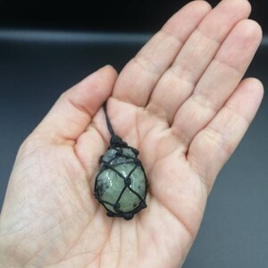 May include: A green crystal stone is wrapped in a black macrame net and hangs from a black cord.