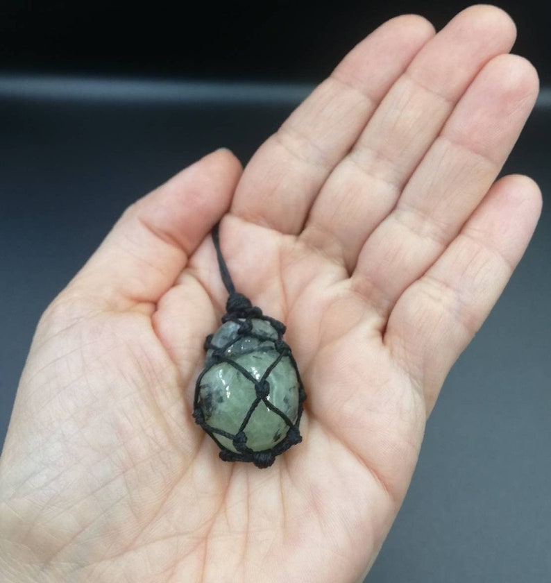 May include: A green crystal stone is wrapped in a black macrame net and hangs from a black cord.
