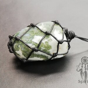 May include: A green and white moss agate stone wrapped in a black macrame cord. The stone is tied with a knot at the top and has a black cord hanging from it.