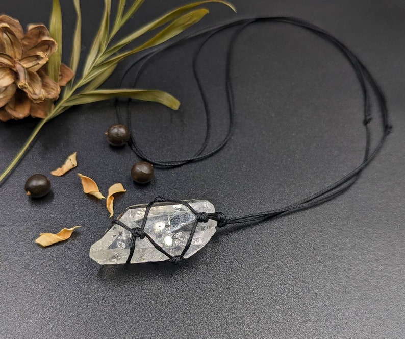 May include: A clear quartz crystal pendant on a black cord necklace. The crystal is wrapped with black cord and has a raw, natural shape.