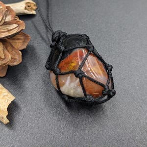 Peut inclure: Une pierre d'agate rouge, orange et blanche enveloppée d'un cordon en macramé noir. La pierre est nouée en haut, créant un pendentif.