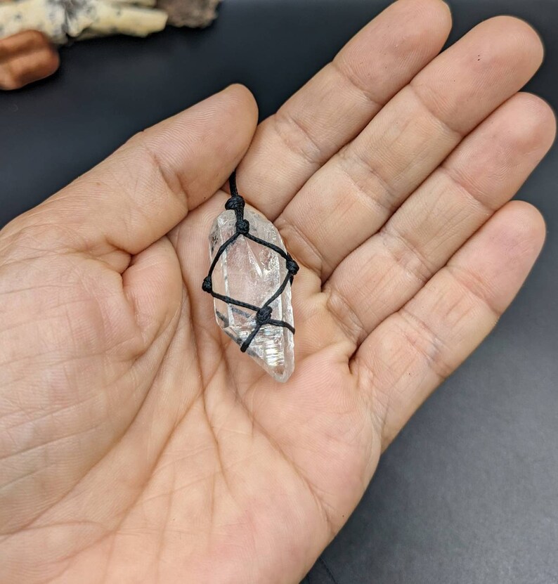 May include: A clear quartz crystal pendant wrapped in black cord. The crystal is faceted and has a natural, raw shape.