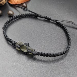 Op de afbeelding: Een armband van zwart gevlochten koord met een groene kristallen hanger.