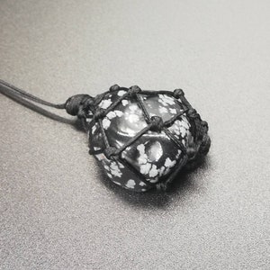 Könnte beinhalten: Ein schwarzer und weißer Schneeflocken-Obsidian-Stein-Anhänger, umwickelt mit schwarzem Kordel und in einem Makramee-Knoten gebunden.