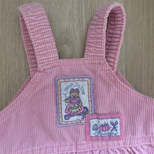 May include: Pale pink corduroy overalls with adjustable straps. Includes two embroidered patches: one with a bear sewing and the text "A Stitch in Time", and another with sewing-themed icons and the brand "Oshkosh".
