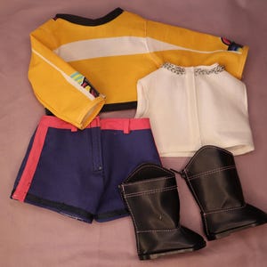 Op de afbeelding: Een poppenoutfitset met een geel en wit gestreept shirt met lange mouwen, marineblauwe shorts met roze rand, een wit vest en zwarte laarzen. De outfit is voor een pop.