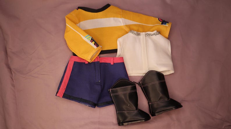 Op de afbeelding: Een poppenoutfitset met een geel en wit gestreept shirt met lange mouwen, marineblauwe shorts met roze rand, een wit vest en zwarte laarzen. De outfit is voor een pop.
