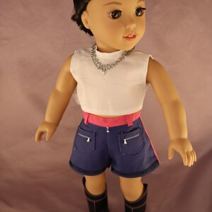 Op de afbeelding: Pop met een witte crop top, blauwe shorts met roze bies en zwarte laarzen. De pop heeft zwart haar met paarse highlights en een zilveren ketting.