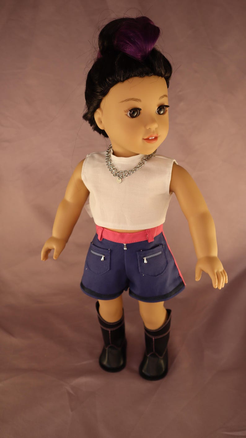 Op de afbeelding: Pop met een witte crop top, blauwe shorts met roze bies en zwarte laarzen. De pop heeft zwart haar met paarse highlights en een zilveren ketting.