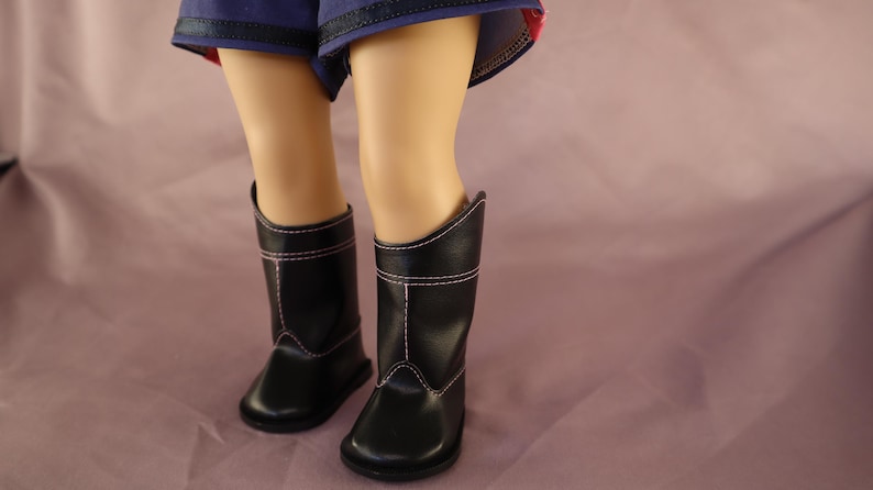 Op de afbeelding: Een paar zwarte poppenlaarzen met witte stiksels. De laarzen hebben een klassiek ontwerp met een ronde neus en een middelhoge hoogte. De laarzen zijn te zien op de benen van een pop, met een blauwe korte broek erboven.