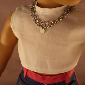 Op de afbeelding: Een pop draagt een witte mouwloze crop top en een zilveren ketting met een kleine ronde hanger. De pop draagt ook een blauwe broek met roze accenten.