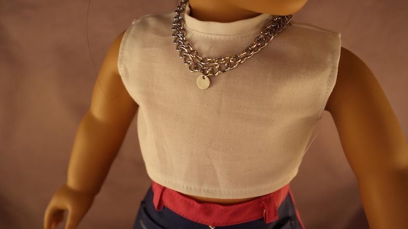 Op de afbeelding: Een pop draagt een witte mouwloze crop top en een zilveren ketting met een kleine ronde hanger. De pop draagt ook een blauwe broek met roze accenten.