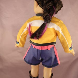 Op de afbeelding: Pop met een gele jas met witte strepen, een blauwe korte broek met roze rand en zwarte laarzen. De pop heeft donker haar in een vlecht met paarse highlights. De pop wordt van achteren bekeken.