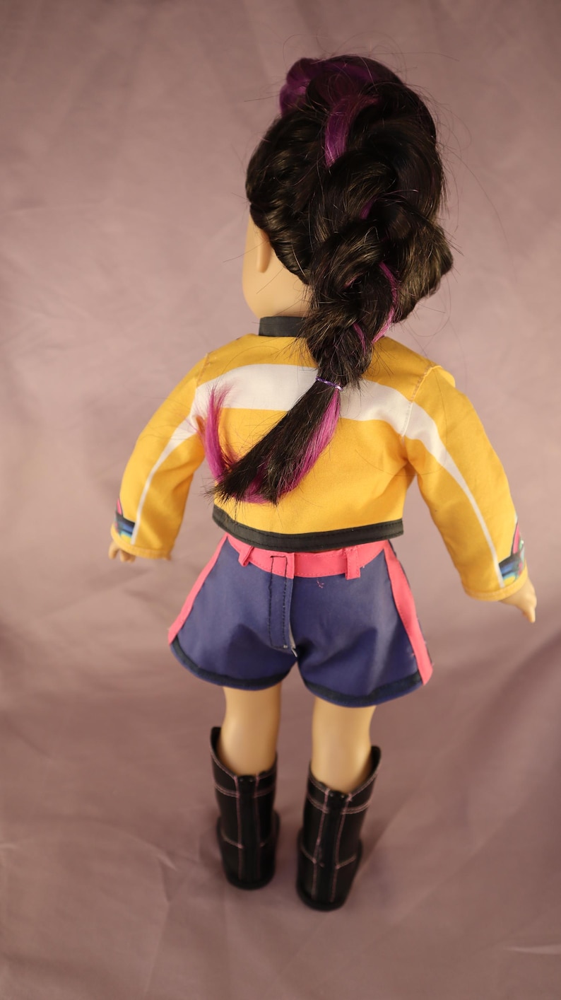 Op de afbeelding: Pop met een gele jas met witte strepen, een blauwe korte broek met roze rand en zwarte laarzen. De pop heeft donker haar in een vlecht met paarse highlights. De pop wordt van achteren bekeken.