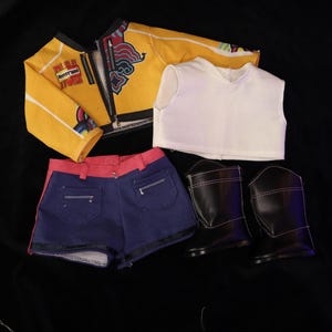 Op de afbeelding: Een poppenoutfitset met een gele jas met rode en witte accenten, een witte mouwloze top, blauwe spijkerbroekshorts met rode bies en zwarte laarzen. De kleding ligt op een zwarte ondergrond.