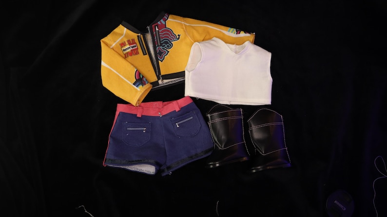 Op de afbeelding: Een poppenoutfitset met een gele jas met rode en witte accenten, een witte mouwloze top, blauwe spijkerbroekshorts met rode bies en zwarte laarzen. De kleding ligt op een zwarte ondergrond.