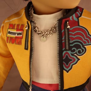 Op de afbeelding: Een pop met een gele jas met zwarte bies en een decoratief ontwerp aan de rechterkant. De jas heeft de tekst "Rrrrrrumi" op de linkermouw. De pop draagt ook een zilveren ketting en een wit shirt.