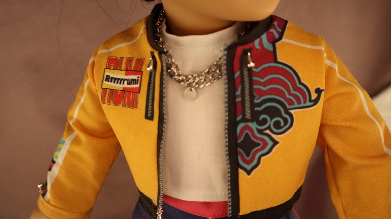 Op de afbeelding: Een pop met een gele jas met zwarte bies en een decoratief ontwerp aan de rechterkant. De jas heeft de tekst "Rrrrrrumi" op de linkermouw. De pop draagt ook een zilveren ketting en een wit shirt.