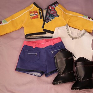 Op de afbeelding: Een poppenoutfit met een gele jas met zwarte rand, een blauwe short met roze rand, een witte top met een zilveren ketting en zwarte laarzen met roze stiksels. De jas heeft een rits.