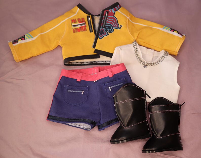 Op de afbeelding: Een poppenoutfit met een gele jas met zwarte rand, een blauwe short met roze rand, een witte top met een zilveren ketting en zwarte laarzen met roze stiksels. De jas heeft een rits.