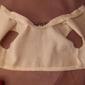 Op de afbeelding: Cr&egrave;mekleurig stoffen vest met armsgaten en een kettingdetail bij de halslijn. Het vest is gemaakt van een zacht, licht materiaal en lijkt handgemaakt. De stiksels zijn zichtbaar langs de randen.