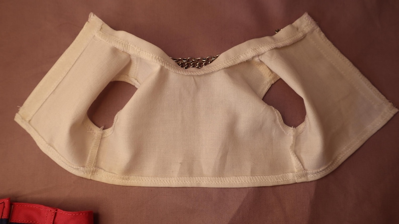 Op de afbeelding: Cr&egrave;mekleurig stoffen vest met armsgaten en een kettingdetail bij de halslijn. Het vest is gemaakt van een zacht, licht materiaal en lijkt handgemaakt. De stiksels zijn zichtbaar langs de randen.