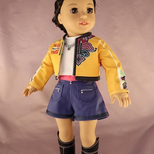 Op de afbeelding: Pop met een gele jas met witte strepen, een wit shirt, blauwe shorts en zwarte laarzen. De jas heeft kleurrijke ontwerpen op de mouwen en de rug. De pop heeft donker haar met een paarse streep.