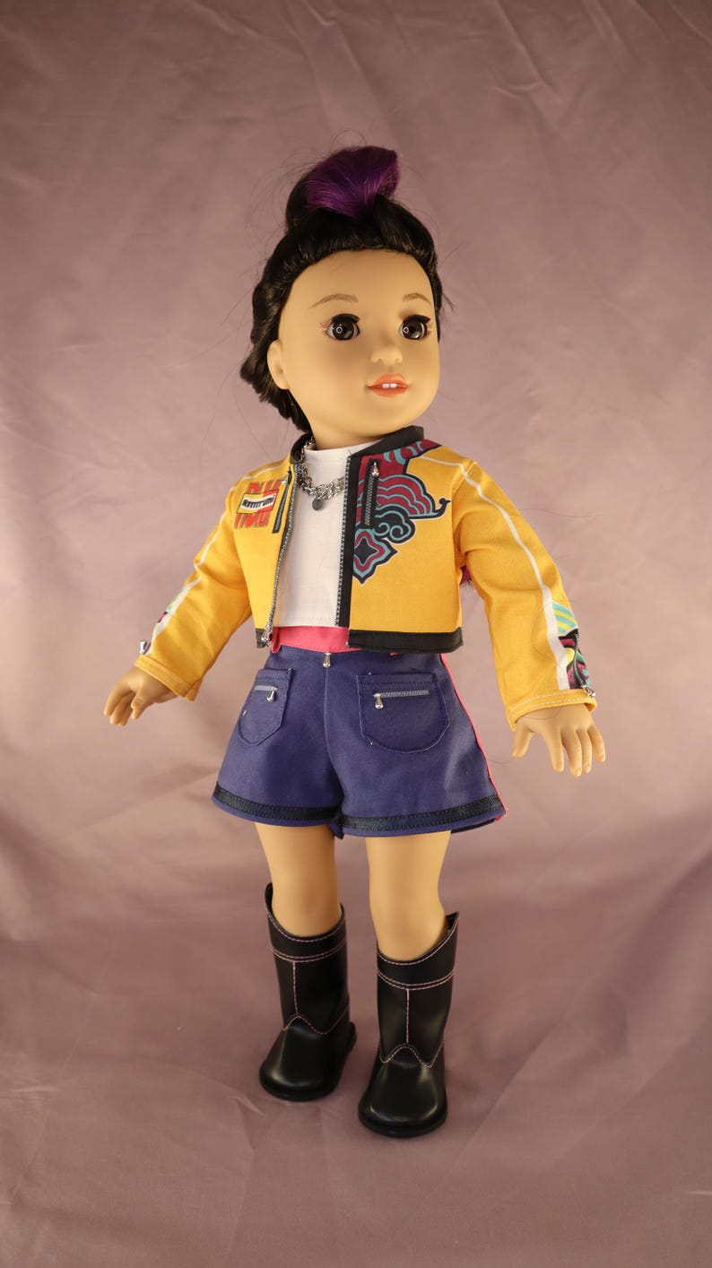 Op de afbeelding: Pop met een gele jas met witte strepen, een wit shirt, blauwe shorts en zwarte laarzen. De jas heeft kleurrijke ontwerpen op de mouwen en de rug. De pop heeft donker haar met een paarse streep.