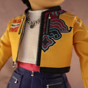 Op de afbeelding: Een pop draagt een gele jas met zwarte afwerking en een decoratief ontwerp aan de voorkant. De jas heeft een ritssluiting en witte strepen op de mouwen. De pop draagt ook een witte top, een zilveren ketting en een blauwe spijkerbroek.